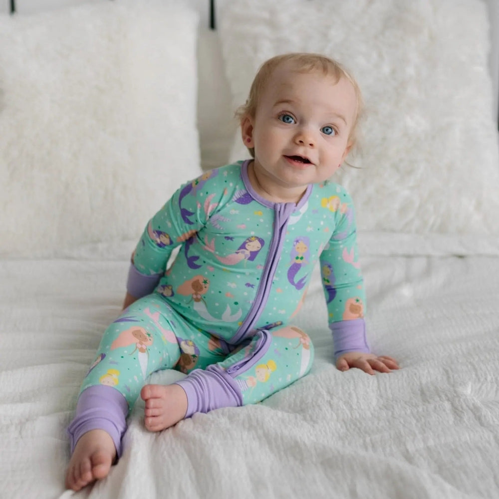 Littlesleepies Mermaid Magic Bamboo Viscose Zippy baby sleeper in aqua teal with purple cuffs, baby seated on bed.