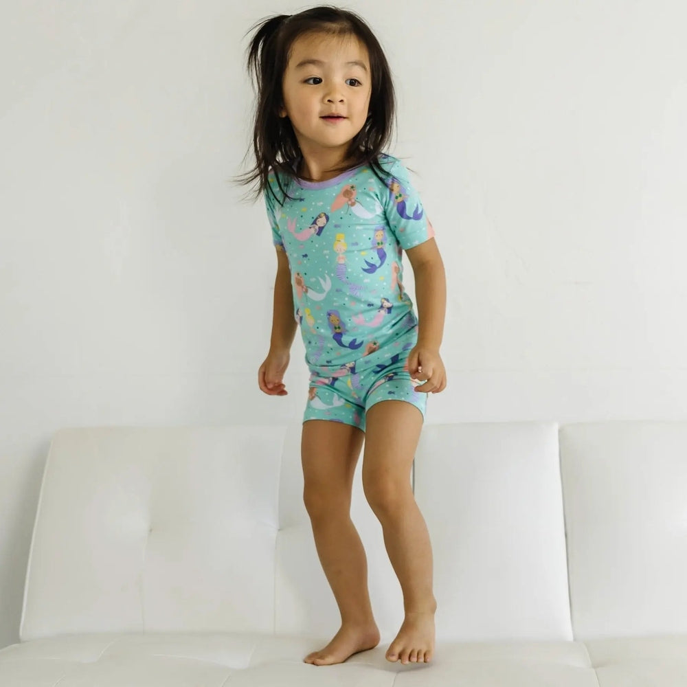 Littlesleepies Mermaid Magic two-piece bamboo viscose pajama set, teal with mermaids, as a child jumps on a white sofa.