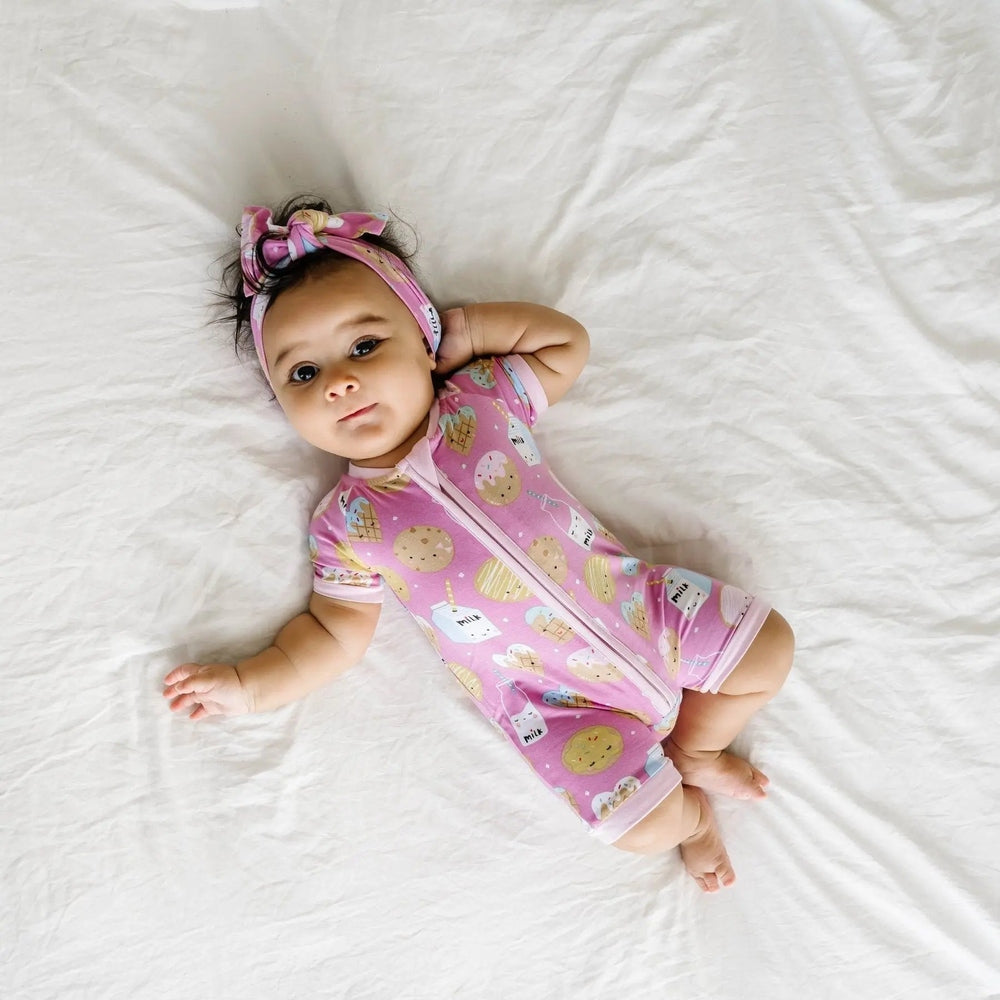 Littlesleepies Pink Cookies & Milk Bamboo Viscose Shorty Zippy: baby in pink cookie-print sleepsuit resting on a white sheet with a headband