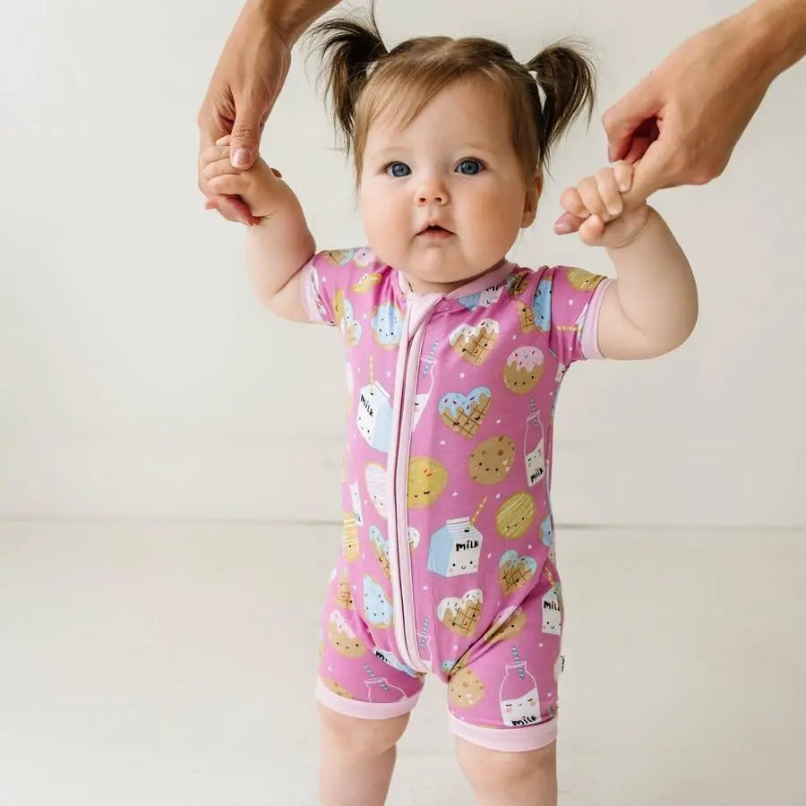 LITTLESLEEPIES Pink Cookies & Milk Bamboo Viscose Shorty Zippy; baby stands in pink sleep romper with a front zipper, held up by adults.