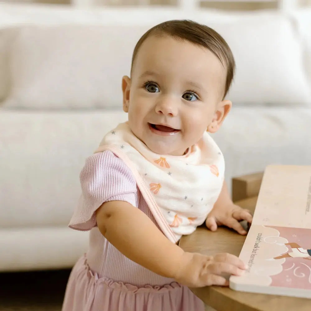 LOULOU LOLLIPOP Bandana Bib Set in pink and white with shell/star print, worn by a smiling baby during feeding.