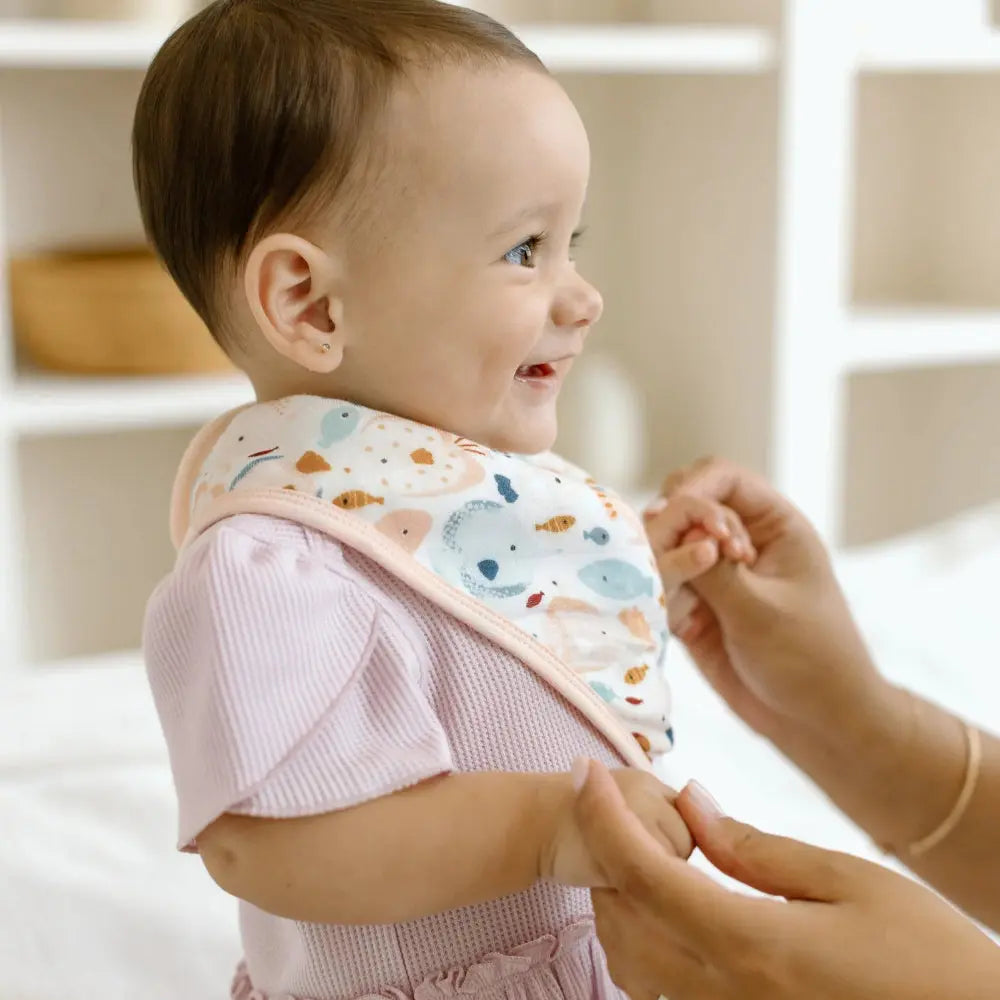 Loulou Lollipop Bandana Bib Set on a smiling baby as caregiver ties the pastel fish-print bib with pink trim.