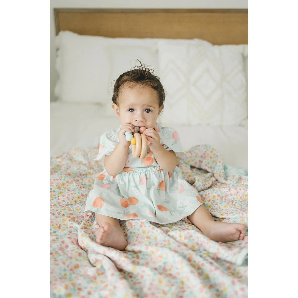 Loulou Lollipop Bodysuit Dress Peaches in pastel mint; baby sits on a floral quilt, chewing peach-toned teether rings.