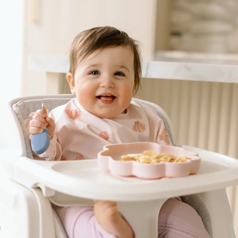 Loulou Lollipop Born To Be Wild Silicone Snack Plate in pink, where a smiling baby scoops snacks from the divided plate.