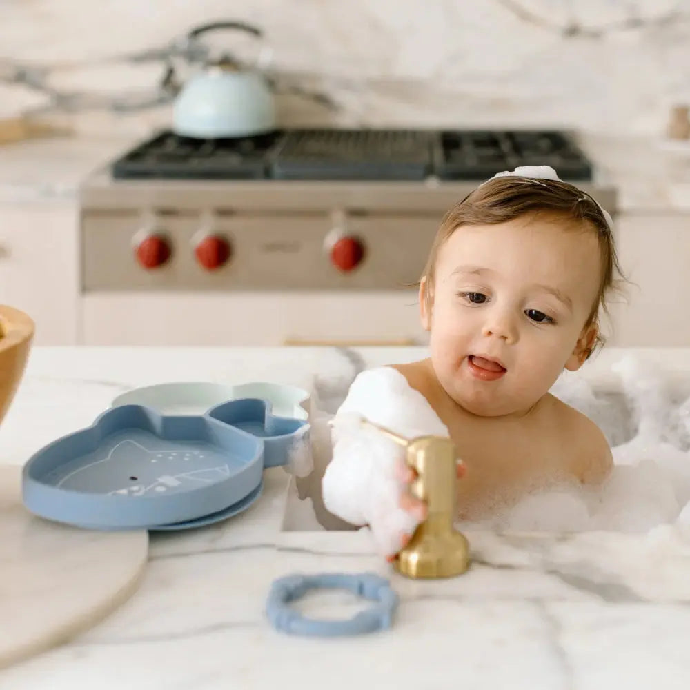 Loulou Lollipop Born To Be Wild silicone snack plate in blue; baby touches its divided compartments while at the bath.