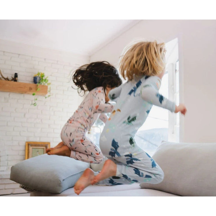 Two kids in LOULOU LOLLIPOP Long Sleeve Pajama Set Magical Dragons leap on a bright couch, showcasing cozy sleepwear.