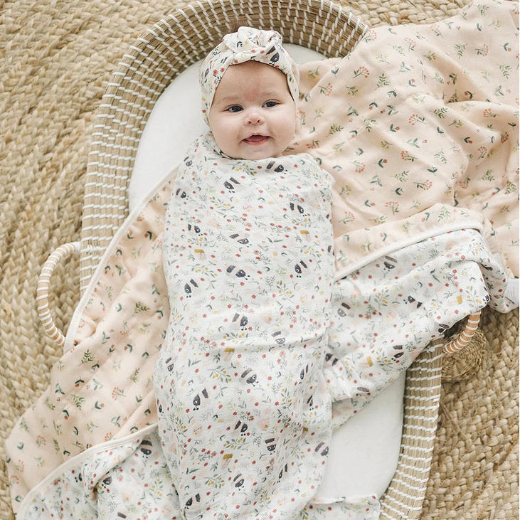 Loulou Lollipop Muslin Quilt Blanket floral print swaddling a smiling baby in a wicker bassinet.
