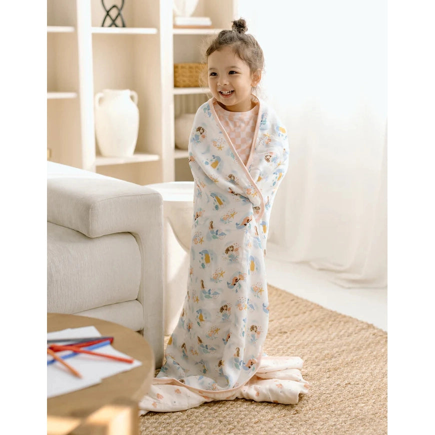 Loulou Lollipop Muslin Quilt Blanket in pastel blue, swaddled around a smiling toddler in a bright living room.