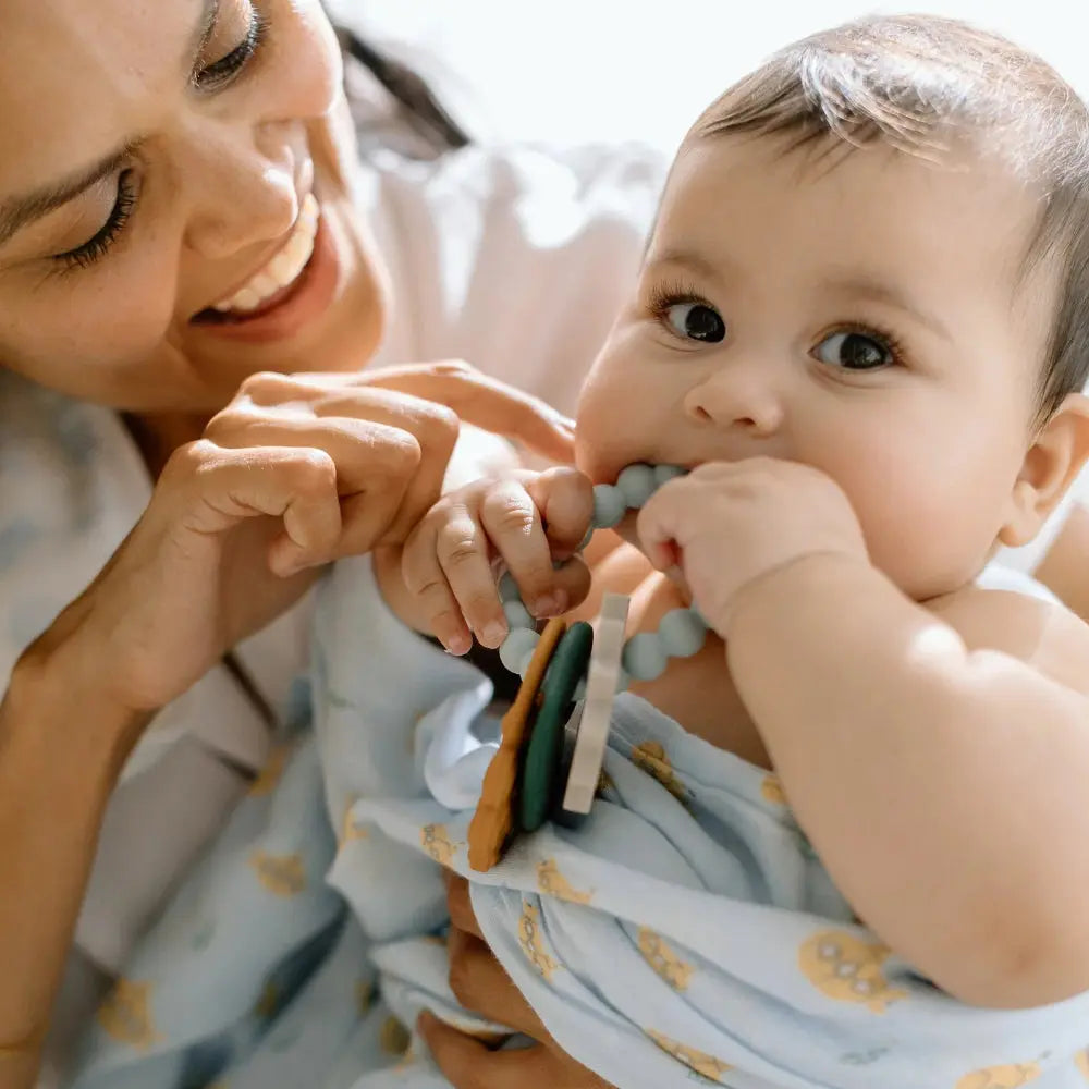 Loulou Lollipop Muslin Swaddle in light blue, baby chewing teething beads while caregiver holds.