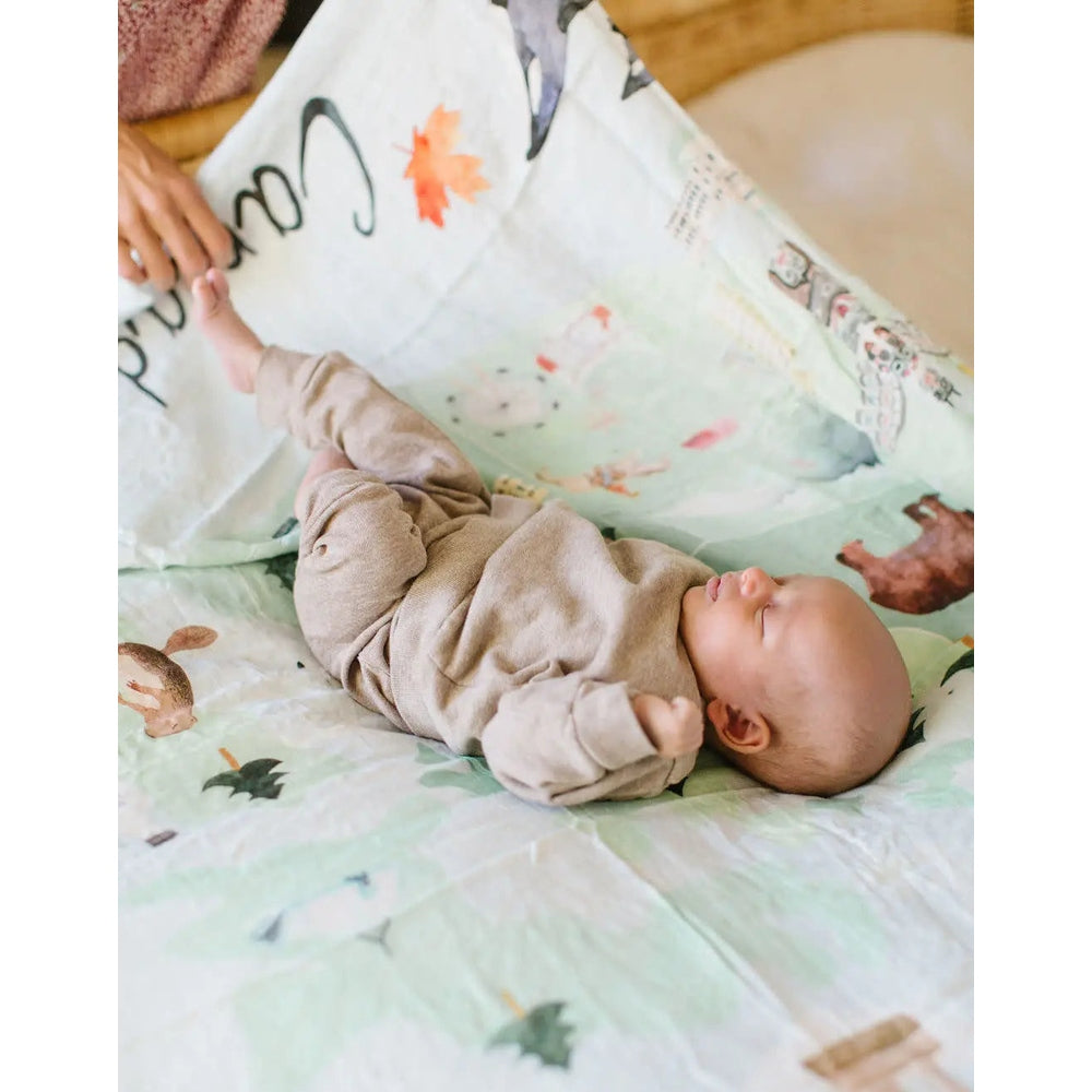 Loulou Lollipop Muslin Swaddle; newborn in beige rests peacefully on pastel-printed blanket.