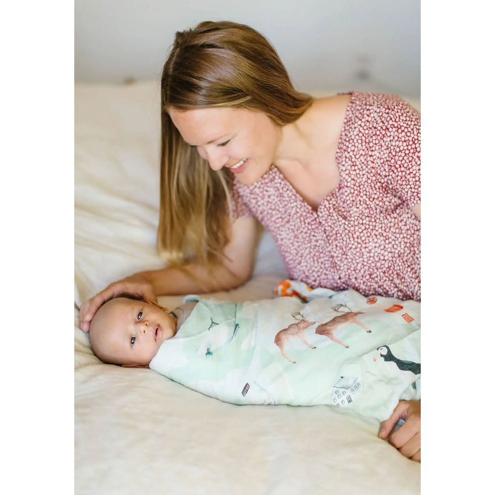 Loulou Lollipop Muslin Swaddle in pale green with animal prints; mother gently comforts newborn on bed.