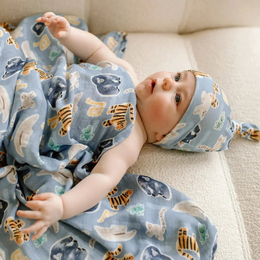 LOULOU LOLLIPOP Muslin Swaddle blanket in blue with tiger prints, baby snugly wrapped on sofa.
