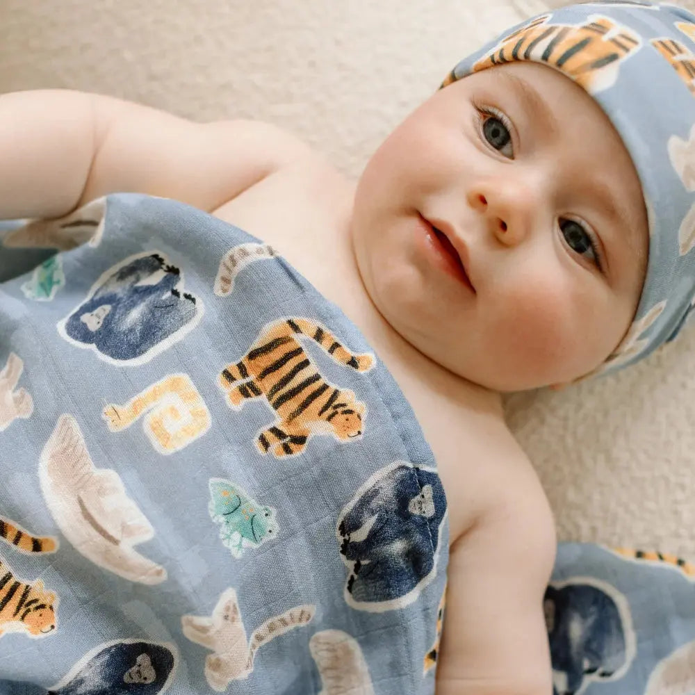 Loulou Lollipop Muslin Swaddle in blue animal print wrapping a smiling baby wearing a matching cap.
