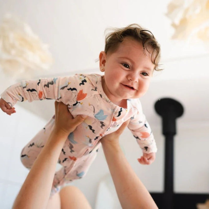 Loulou Lollipop Sleeper Unicorn Ballet, pastel pink baby sleepwear, lifted midair by smiling caregiver.