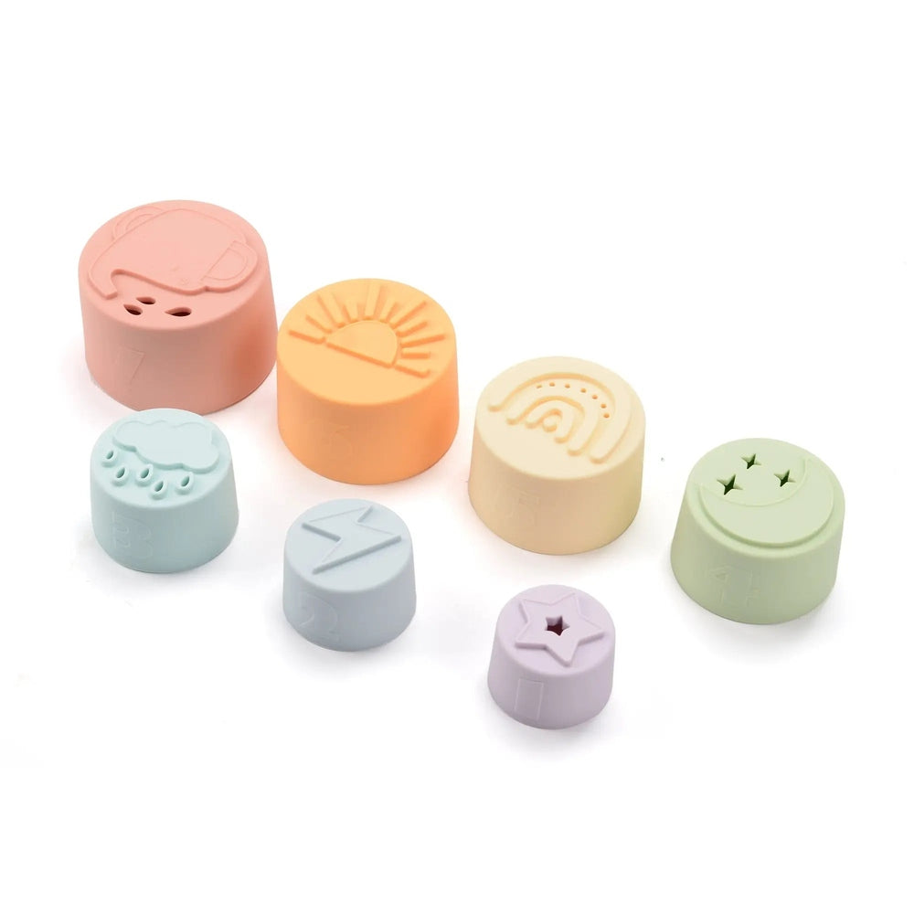 Loulou Lollipop Stacking Cups, pastel silicone 7‑piece set, shown in a row to reveal sun, rainbow, cloud, star patterns.