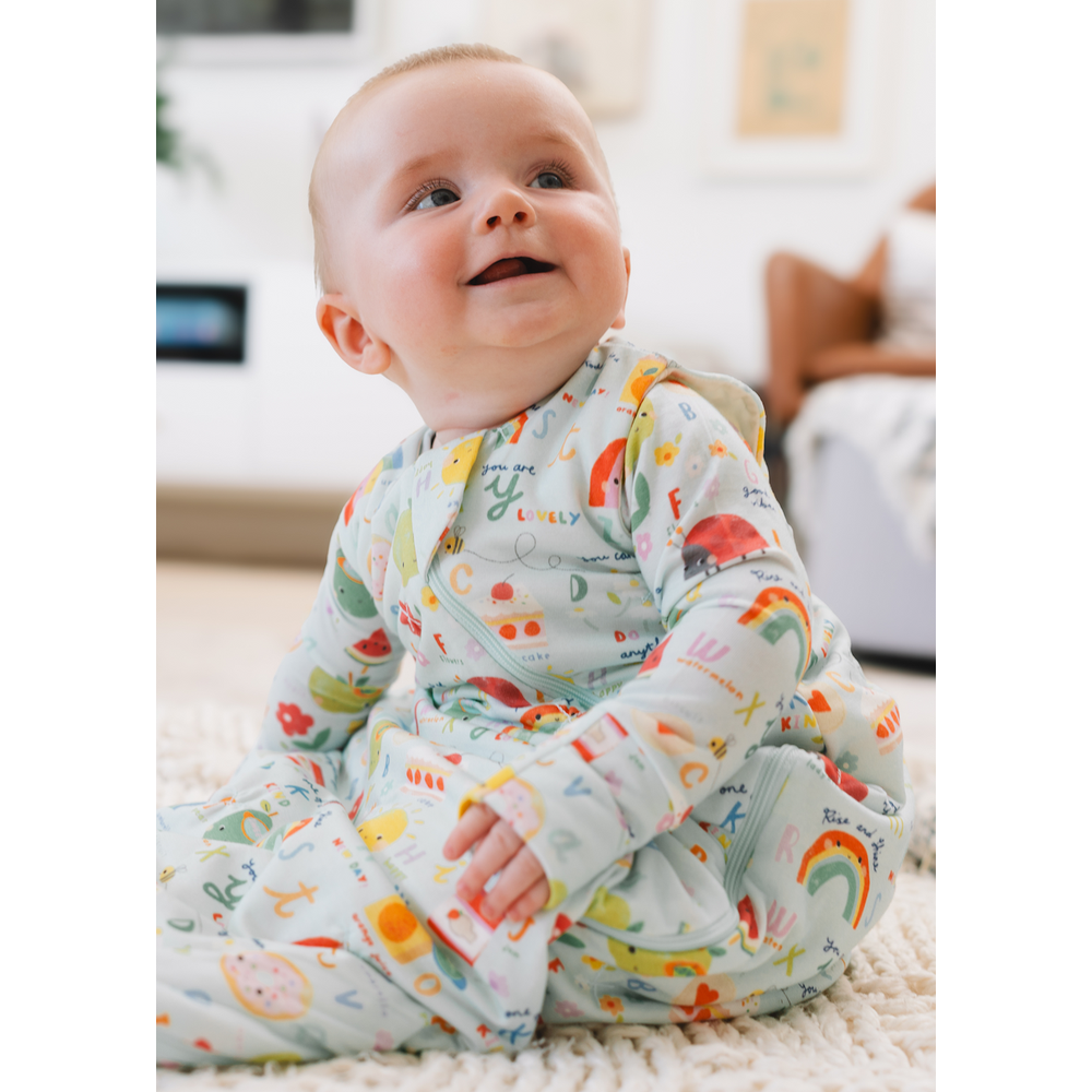 Loulou Lollipop TENCEL Sleep Bag 1.0 TOG Alphabet Smiles; baby sits on a rug, smiling in pastel-print sleepwear.