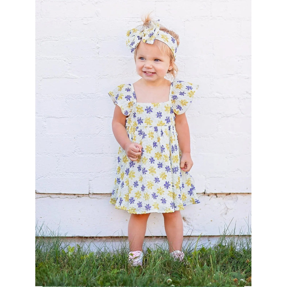 Mabel And Honey Floral Textured Crepe Dress White toddler standing outdoors in a yellow-purple floral dress with matching headband
