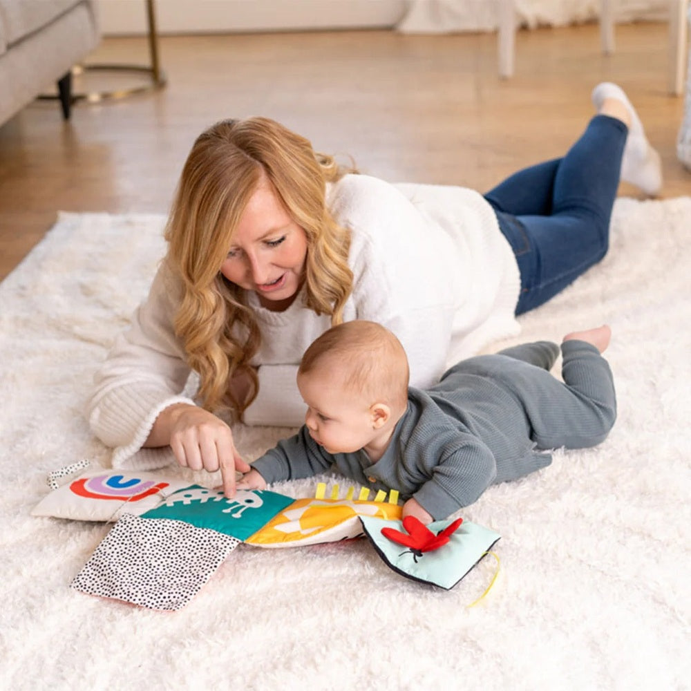 Manhattan Toy Wimmer Seek & Peek Book; mother and baby explore colorful fabric pages on a fluffy rug.