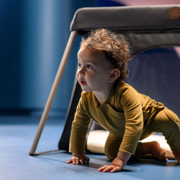 Maxi Cosi Calao travel crib with gray fabric; toddler crawling underneath.