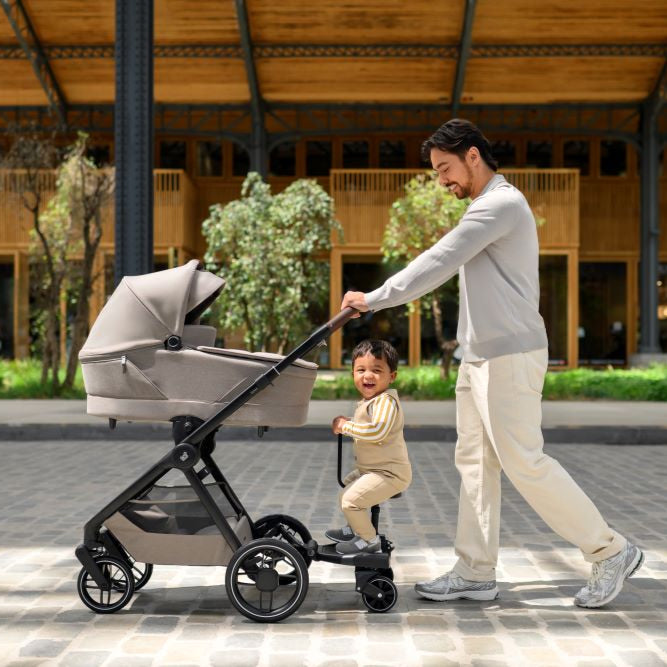 MAXI COSI Hop-on Board attached to a gray stroller; dad guiding toddler on the board during a stroll.