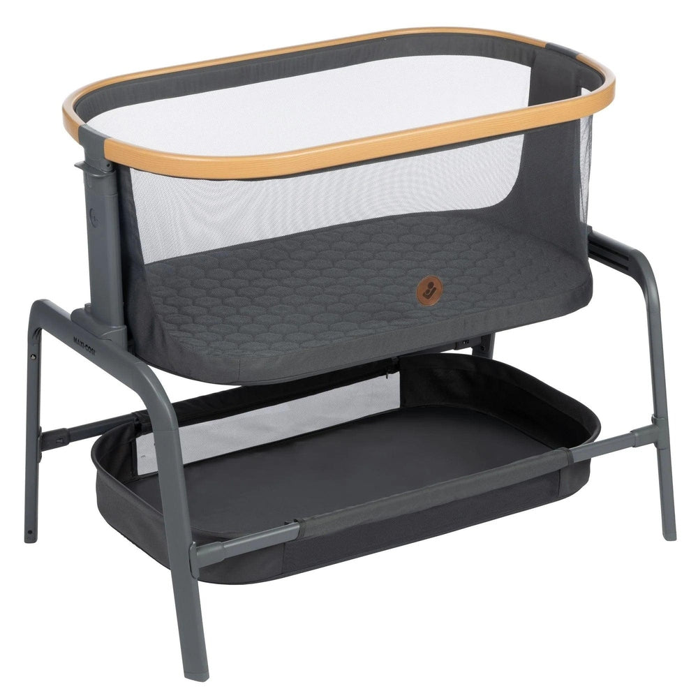 MAXI COSI Iora bedside bassinet in gray with wood trim, mesh sides, and lower storage basket.
