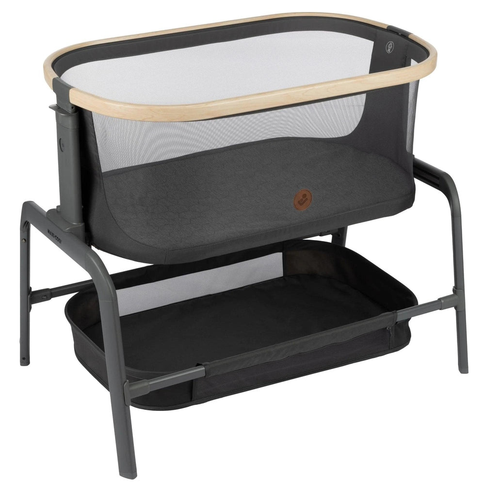 MAXI COSI Iora bedside bassinet in gray fabric with wood rim and mesh sides, showing a lower storage basket.