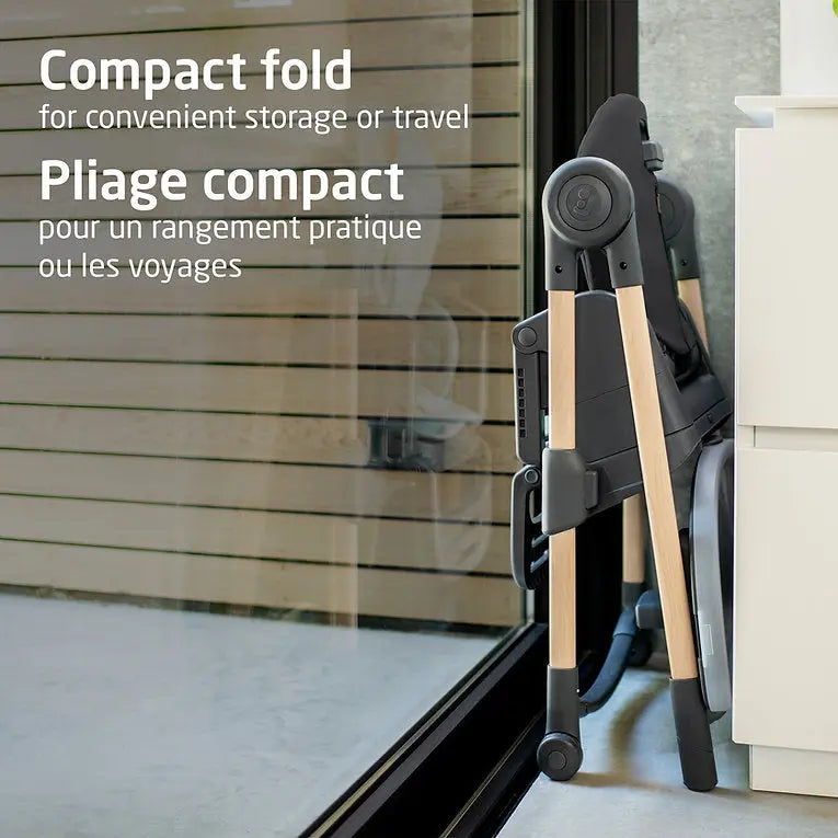 MAXI-COSI Minla 6-in-1 high chair in compact fold with wood-tone legs beside a glass door.