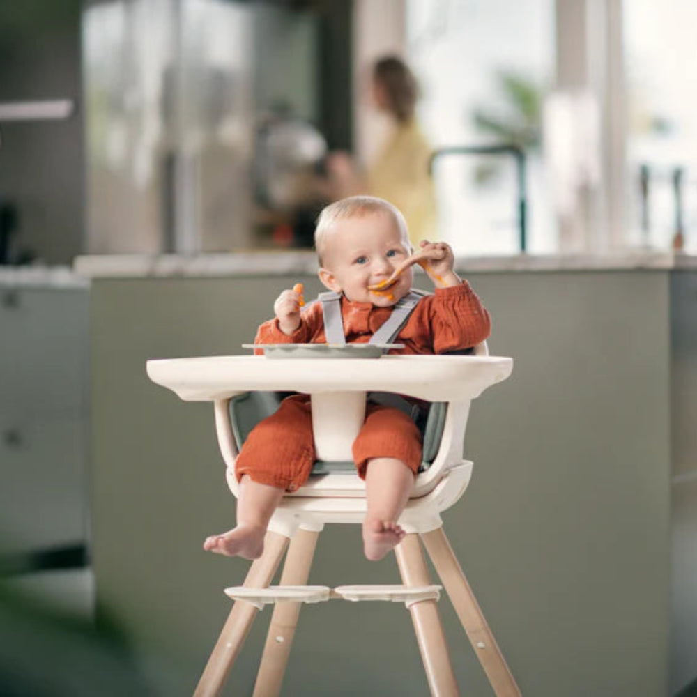 MAXI-COSI Moa 8-in-1 high chair with white tray and wooden legs; baby in orange outfit eating.
