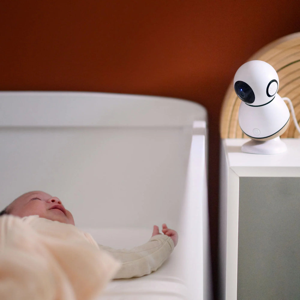 MAXI-COSI See Pro 360° baby monitor on a white nightstand beside a sleeping baby in a bassinet