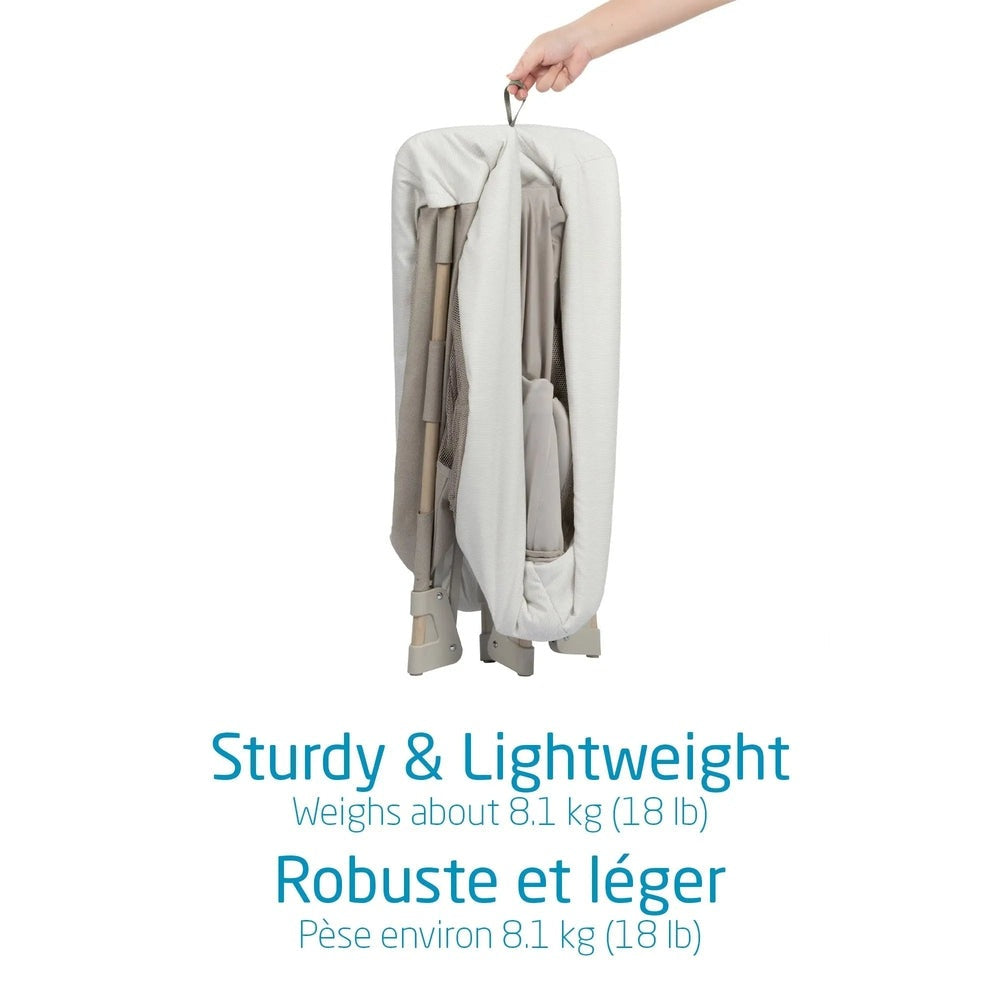 Maxi Cosi Swift Playard folded for easy storage in light gray fabric, hand gripping the carrying handle.