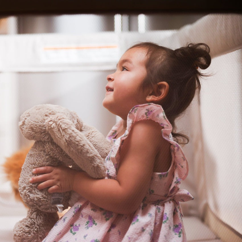 Maxi Cosi Swift Playard in a nursery; toddler in pink floral dress holding a plush stuffed animal