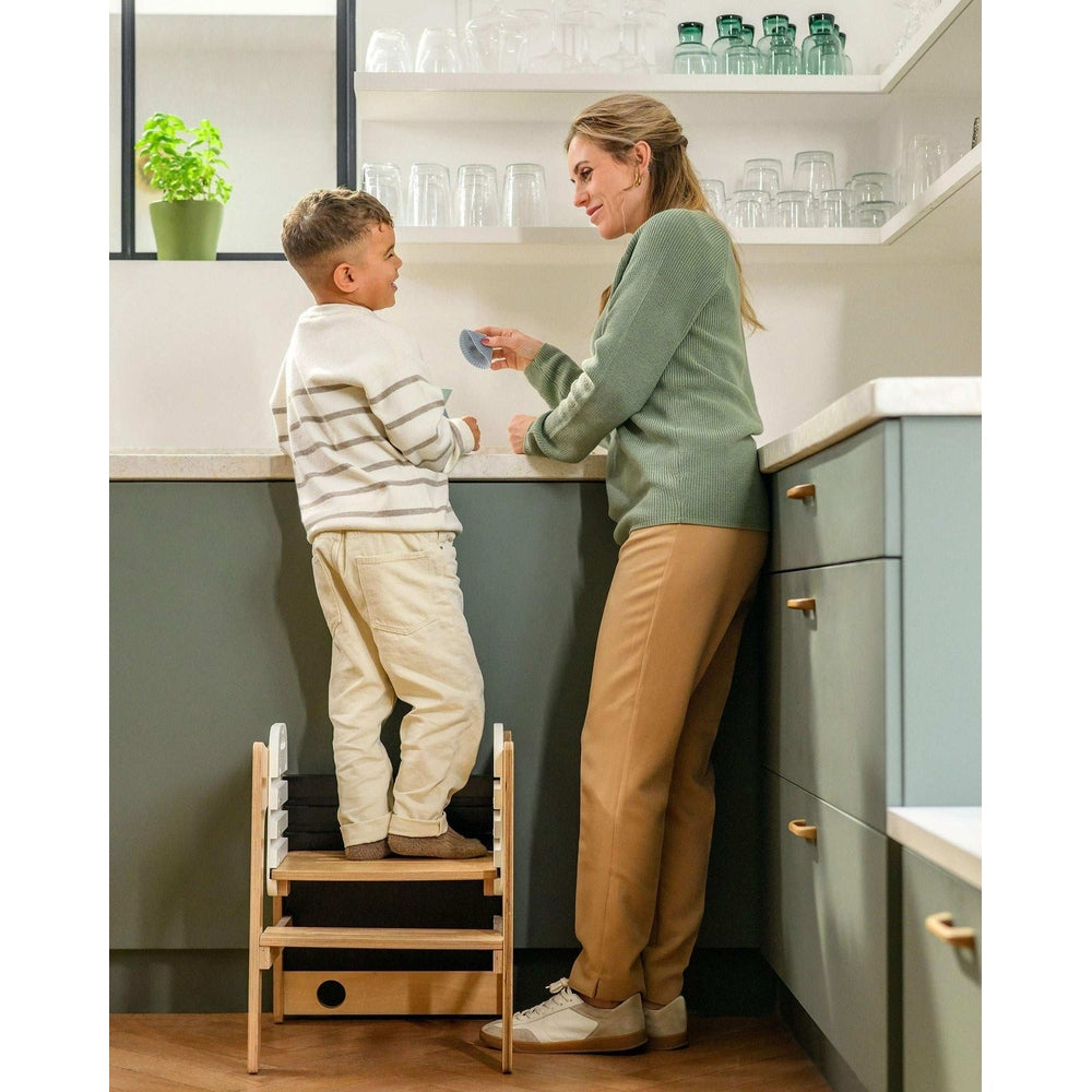 Maxi-Cosi Toucan 3-in-1 learning tower in light wood with gray steps; toddler at the kitchen counter with parent