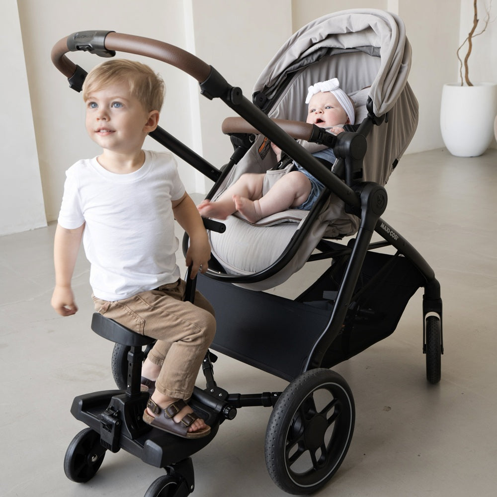 Maxi-Cosi Zelia Luxe 5-In-1 Modular Travel System stroller in beige/gray with a toddler on the front footboard pushing beside a baby in the seat.