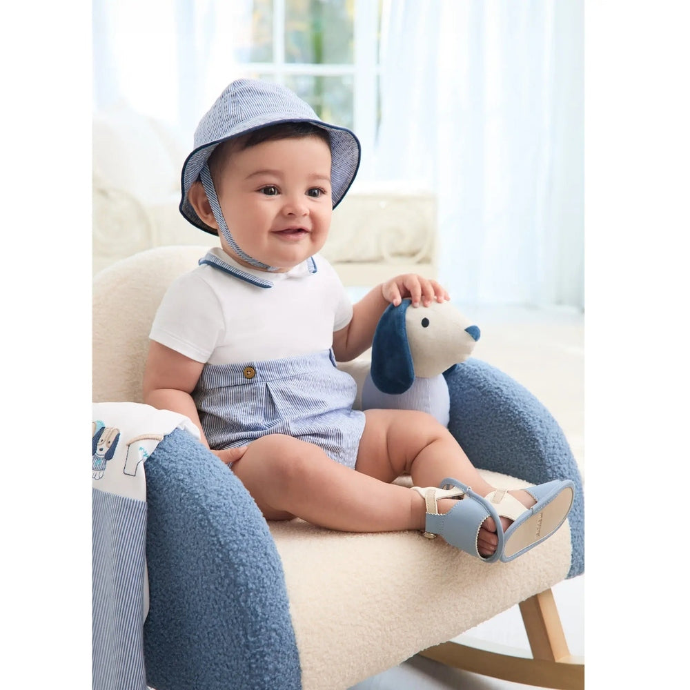 MAYORAL Newborn Romper With Hat Seersucker: baby sits in a plush chair in blue seersucker romper and hat, petting a plush dog.