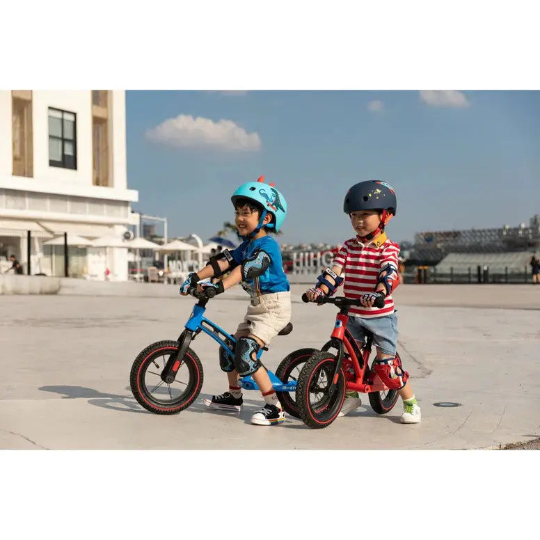 Micro Helmet V2 Scootersaurus: two kids wear blue and red helmets while riding blue and red balance scooters outdoors.