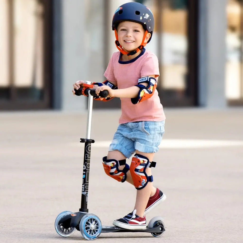 Micro Knee & Elbow Pad Set Black worn by a smiling child on a scooter, showing knee and elbow protection.