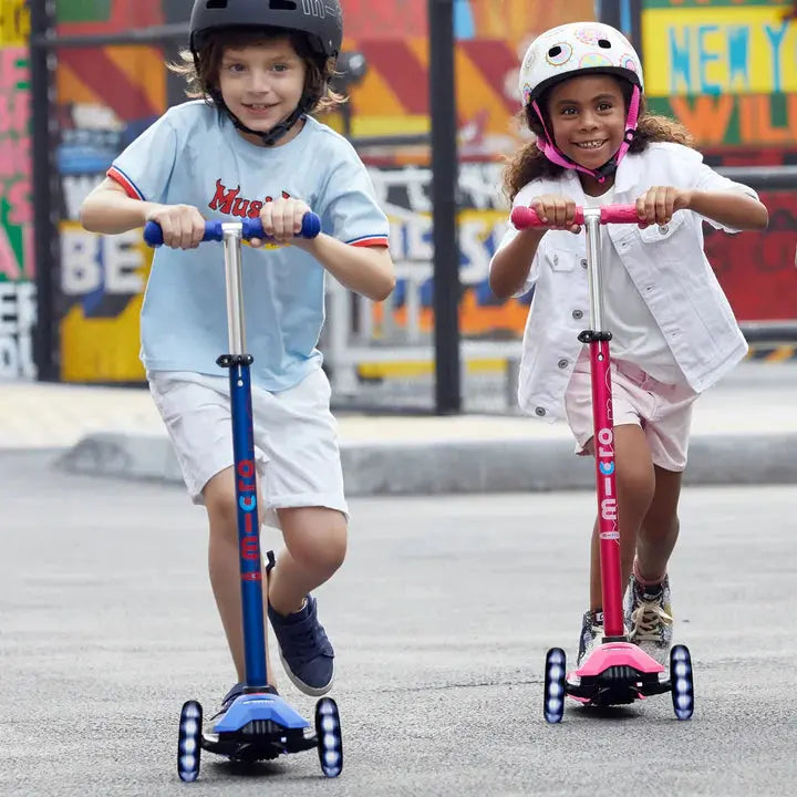 Micro Maxi Deluxe LED Scooter, blue and pink, as two kids ride side-by-side in helmets.