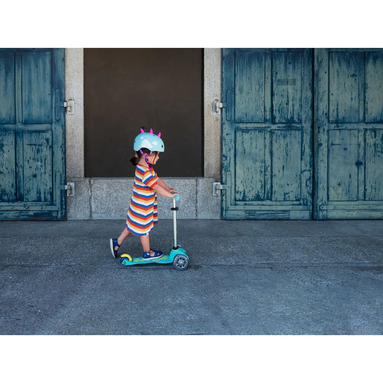 Micro Mini Deluxe Eco Scooter Green; child rides outdoors in a striped dress with a blue helmet.