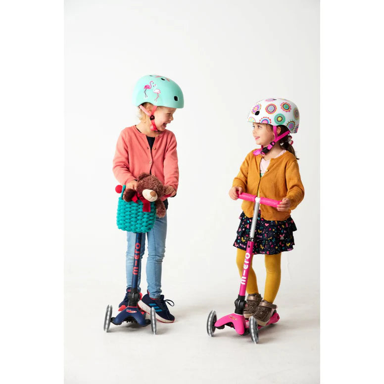 Micro Mini Deluxe Foldable LED Scooter; two kids ride pink and blue scooters in helmets, one with a teddy in a turquoise basket.