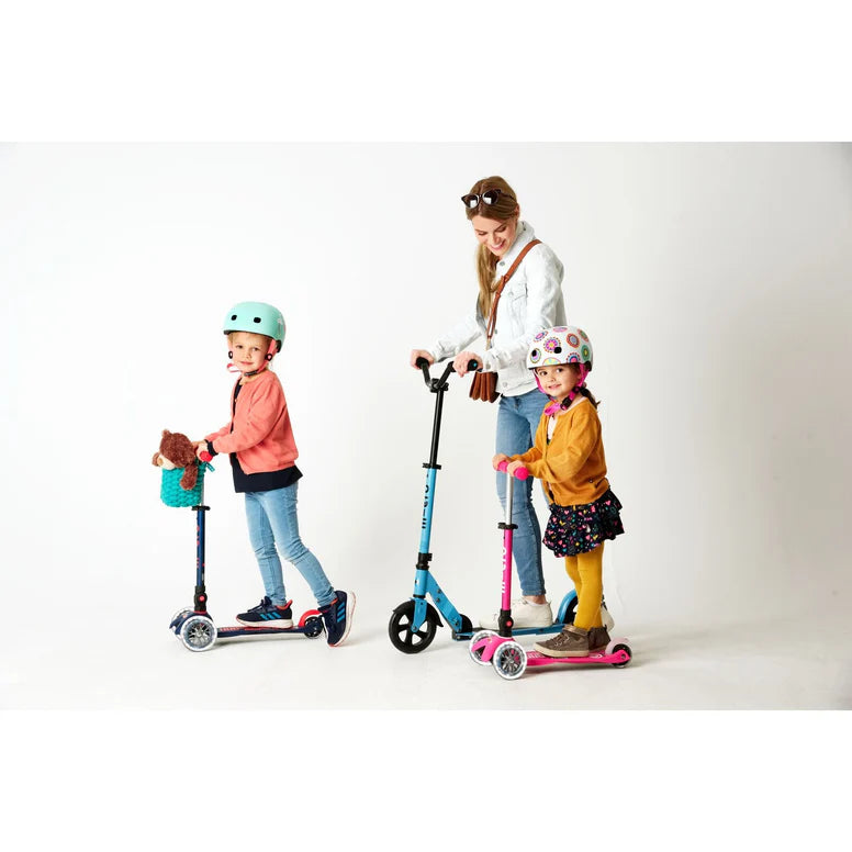 Micro Mini Deluxe Foldable LED Scooter; children ride blue and pink scooters with an adult supervising, helmets on.