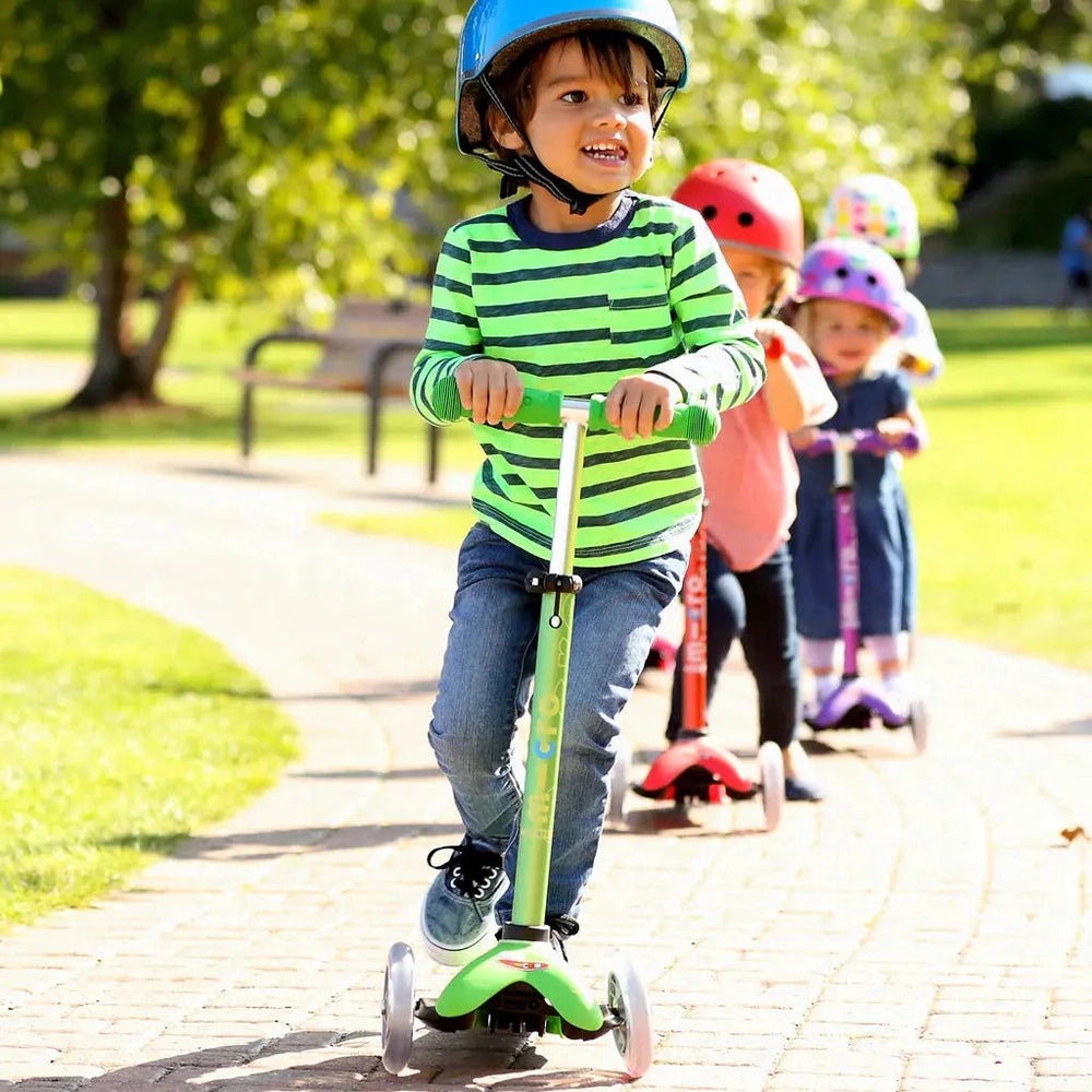 Micro Mini Deluxe Scooter: a child in a blue helmet rides a green scooter along a sunny park path.