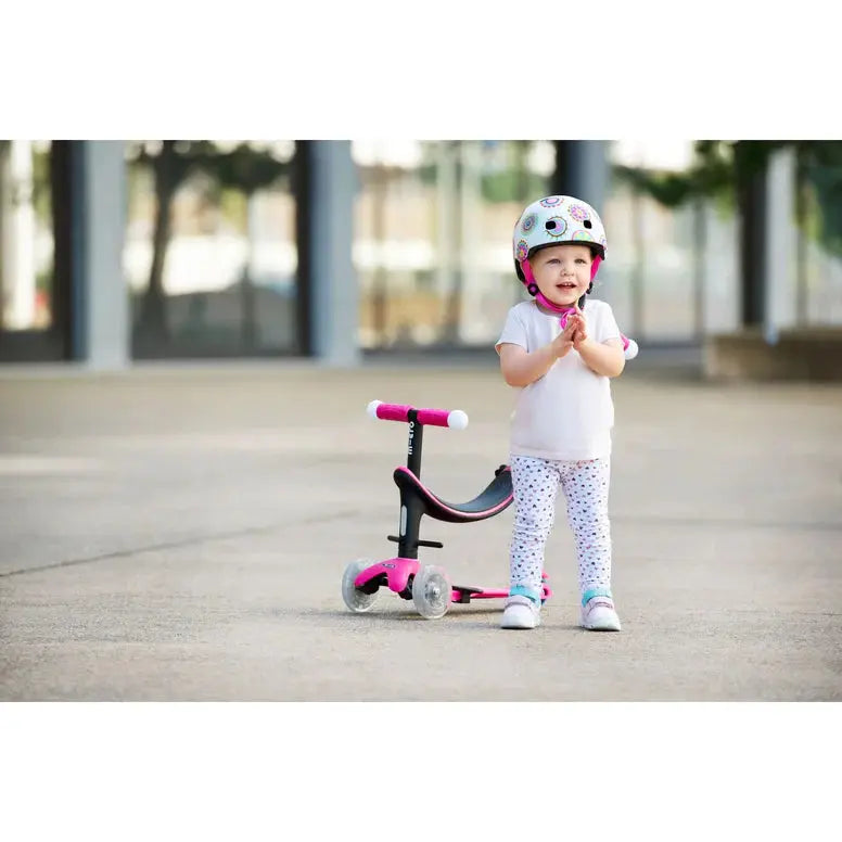 Micro Mini2Grow Deluxe LED Scooter for toddlers; pink accents, three-wheel stability, child stands beside wearing a helmet.