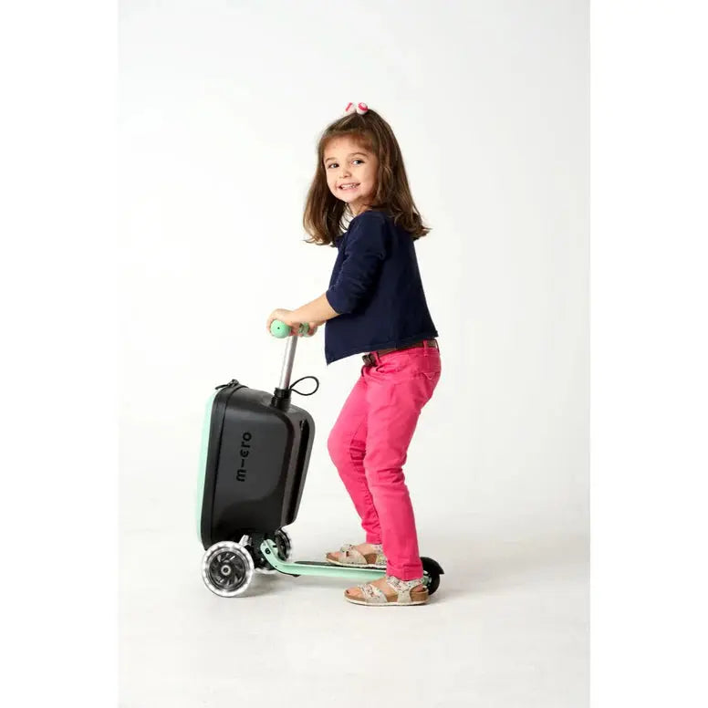 Micro Scooter Junior Luggage Mint suitcase on wheels with mint accents, being ridden by a smiling girl.