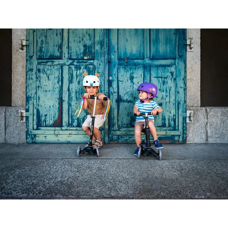 Micro Scooter Streamers: two kids ride black scooters with rainbow streamers, wearing helmets by weathered blue doors.