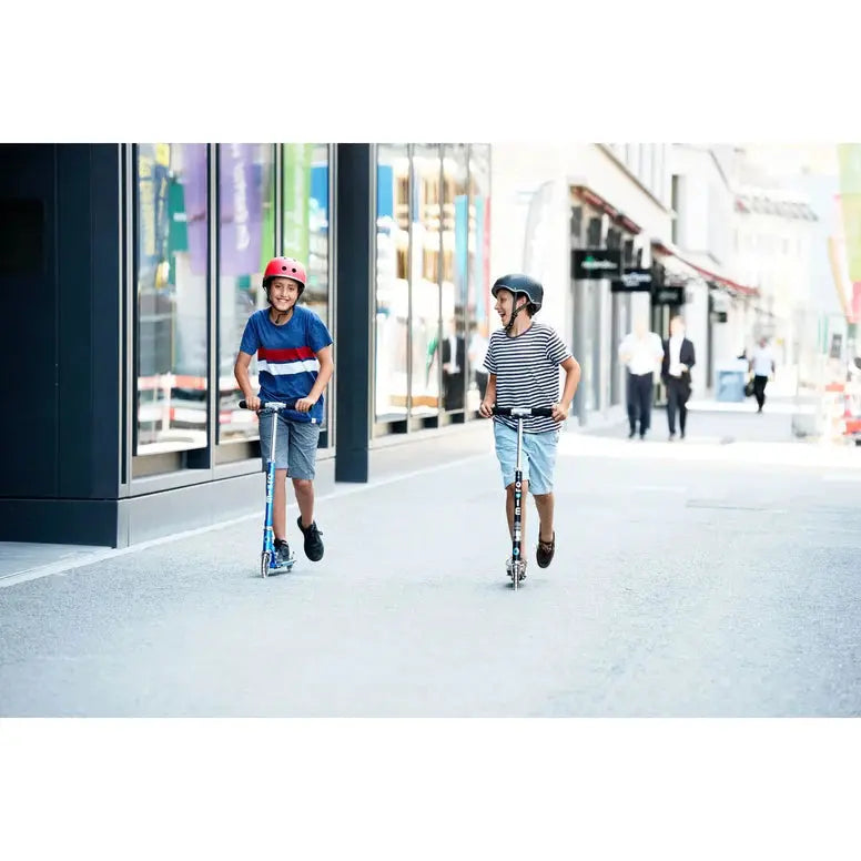 Micro Sprite LED Scooter: two kids wearing helmets ride blue scooters along a busy city street.