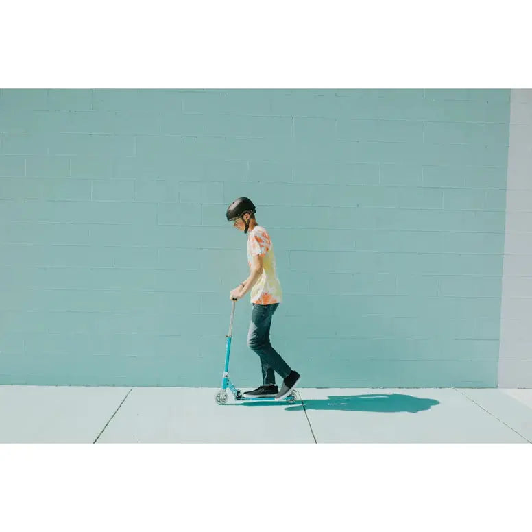 Micro Sprite LED Scooter, turquoise frame, ridden by a person in a helmet along a sunlit sidewalk.