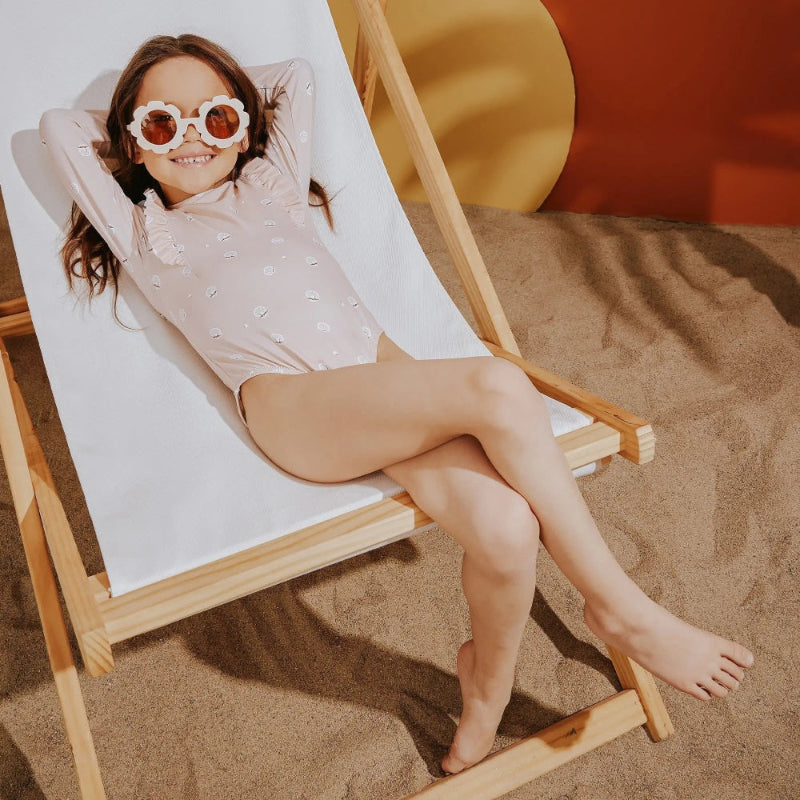 Miles The Label Long Sleeve Swimsuit Pink: a girl lounges on a beach chair, wearing oversized flower sunglasses.