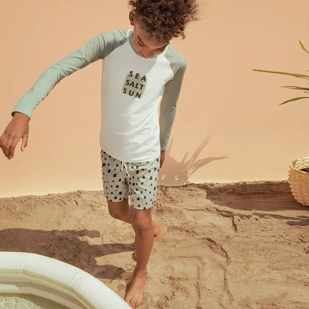 Miles The Label Sea Salt Sun Long-Sleeve Raglan Rashguard dusty green sleeves, white torso, kid stepping by pool edge