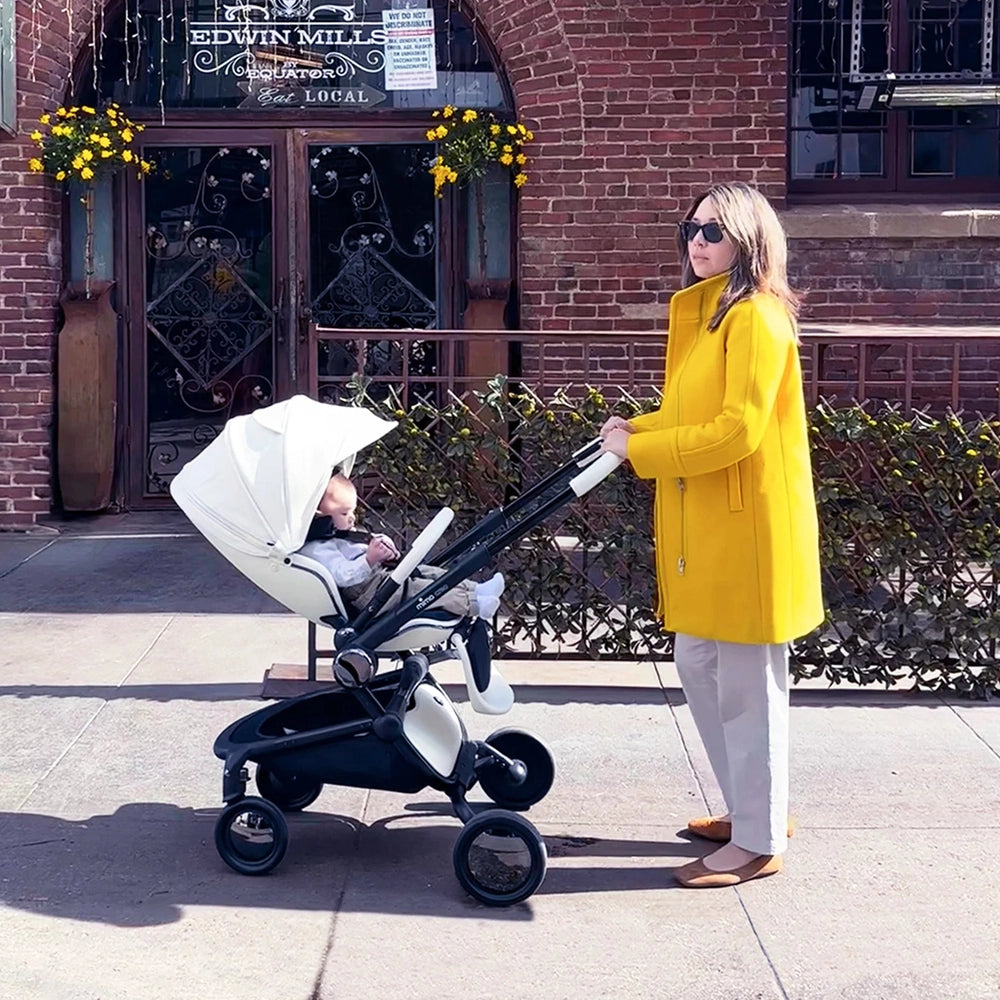 Mima Creo stroller in cream with white canopy being pushed by a woman in a bright yellow coat.
