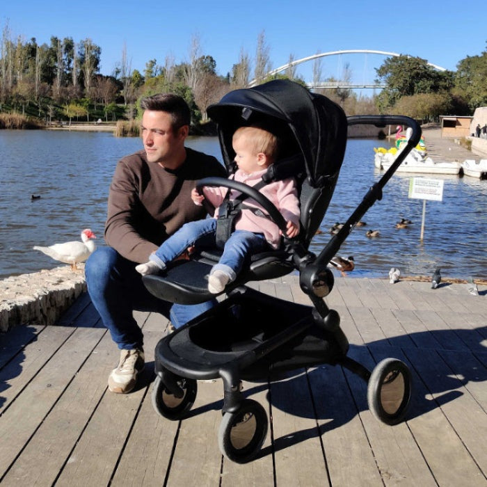 Mima Creo Stroller—black frame with a seated child secured in harness on a lakeside boardwalk.